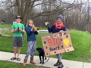 An adult woman, two tweens, and a cute black dog play music while holding a sign that says "Little City, Big Heart."